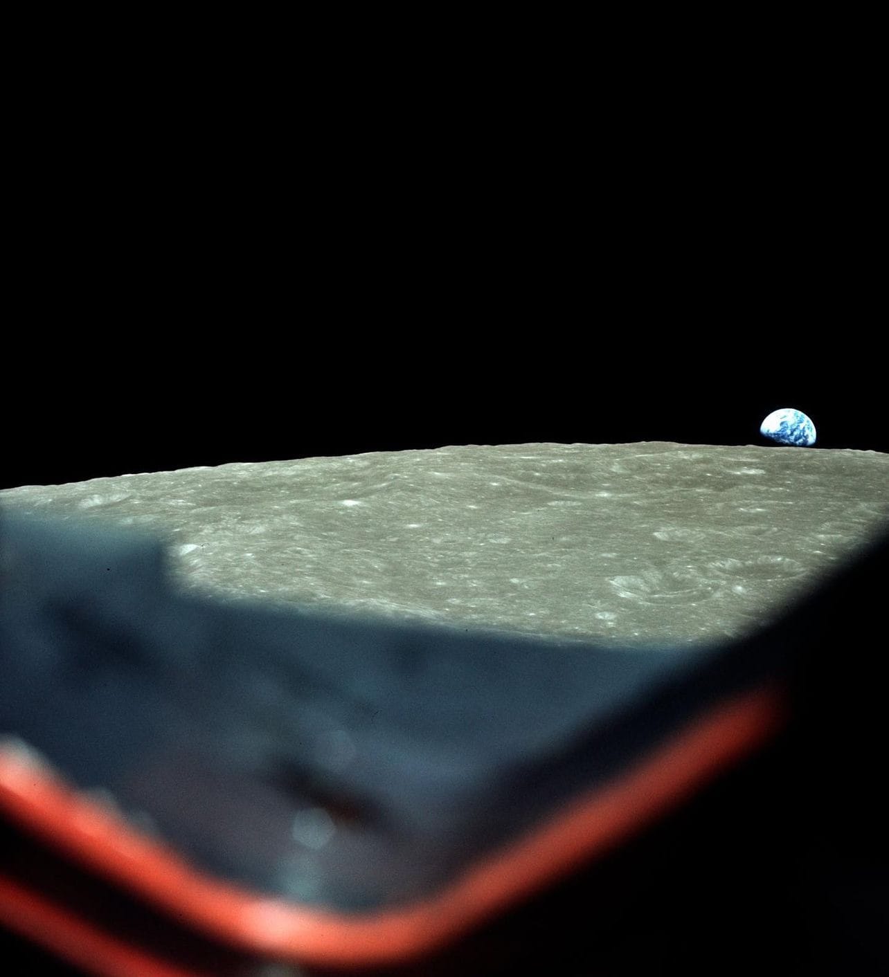Earth rises above the gray lunar surface against the blackness of space, viewed from a spacecraft window that frames the foreground in soft shadow and blurred red edging.